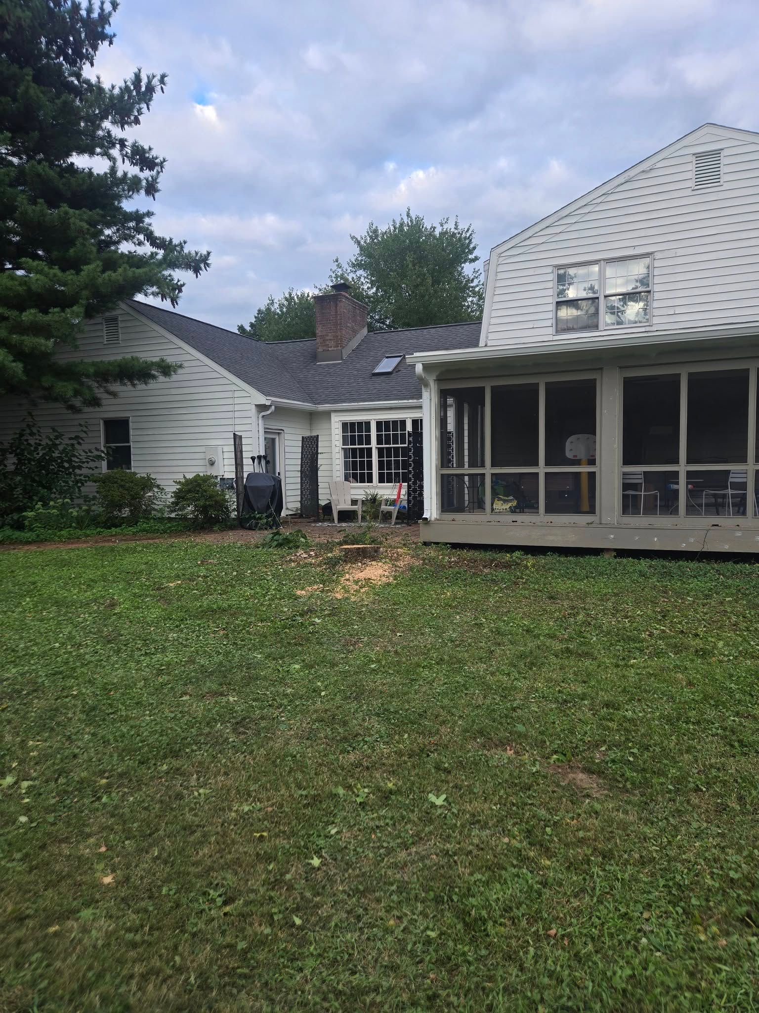 Backyard view: white house and screened porch on the right. Green grass, trees, and cloudy sky.