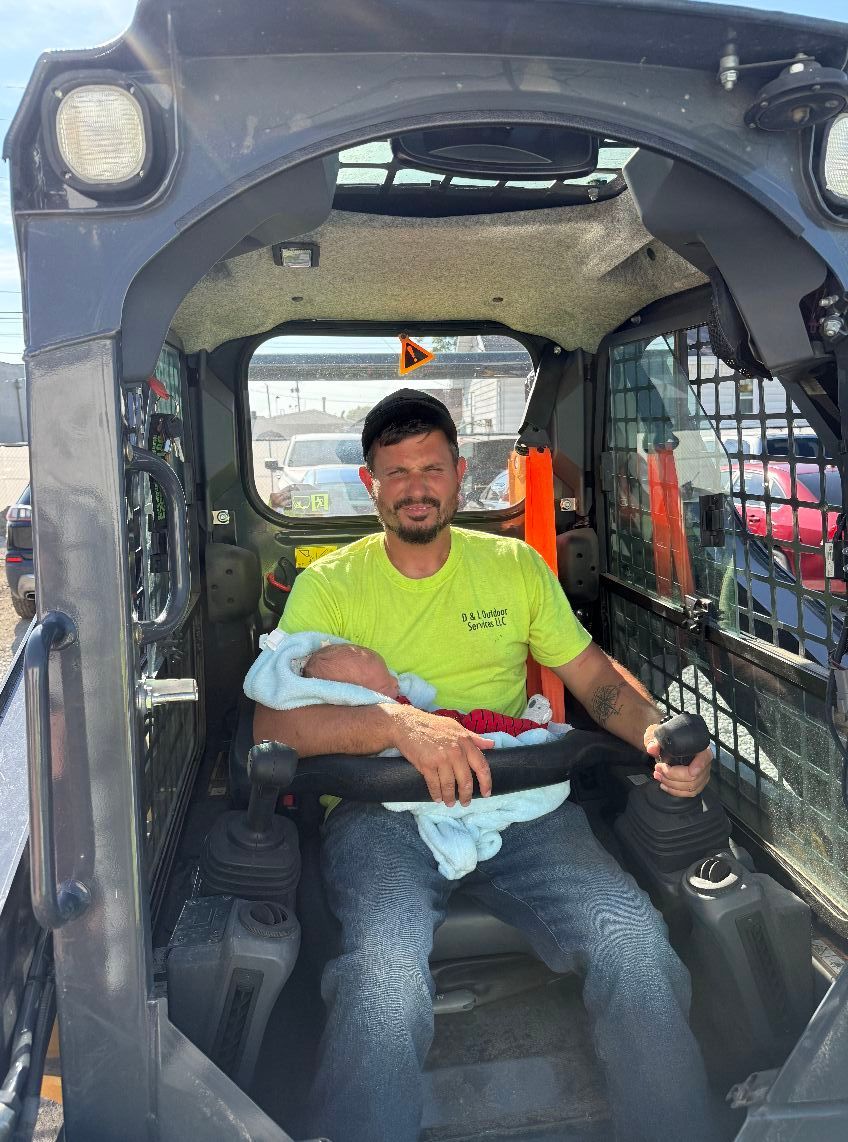 Man in a construction vehicle holding a baby; both in a cab, sunny outdoors.