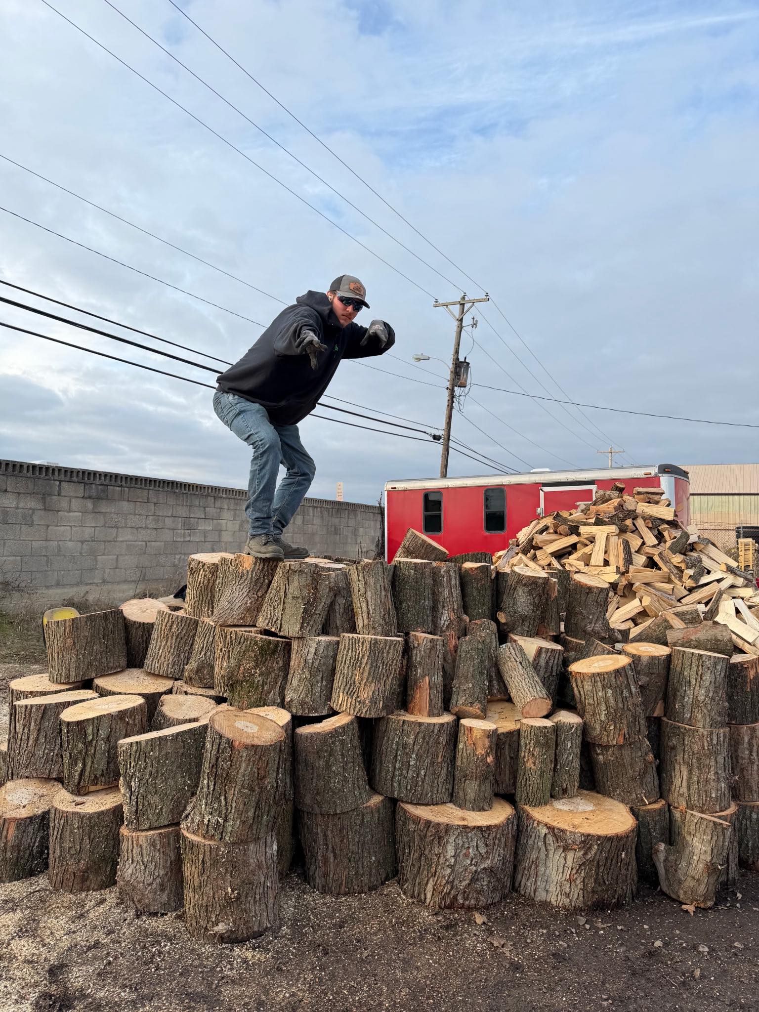 Man balances on a pile of firewood outdoors, reaching towards the camera. Red building and cloudy sky in background.