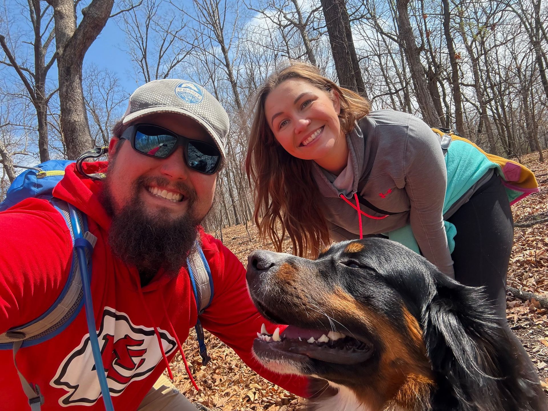 A man in a red hoodie and a woman in a grey zip-up hike with their tricolor dog in a sunny, wooded outdoor setting.