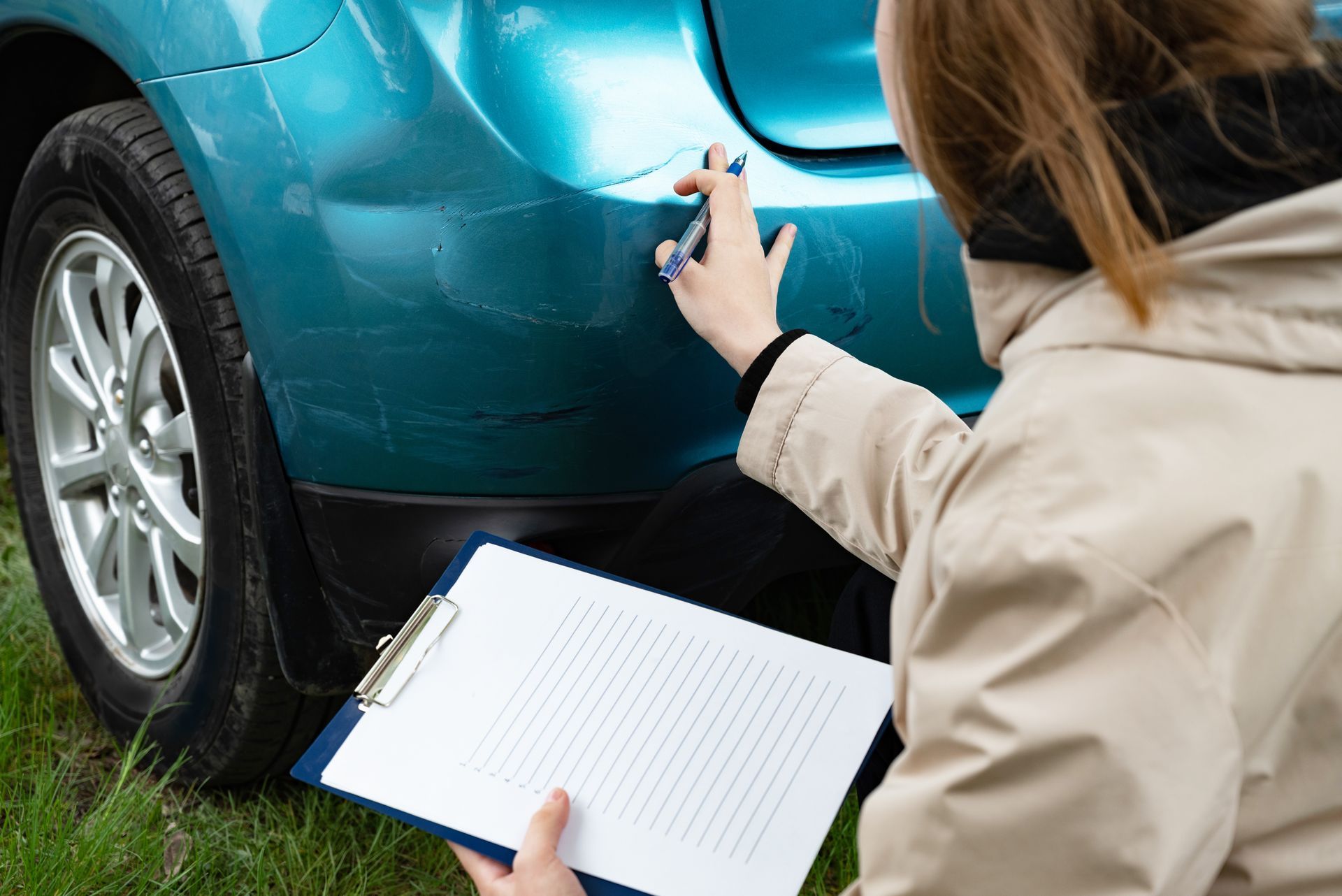 Person inspecting a teal car’s rear bumper with a clipboard outdoors