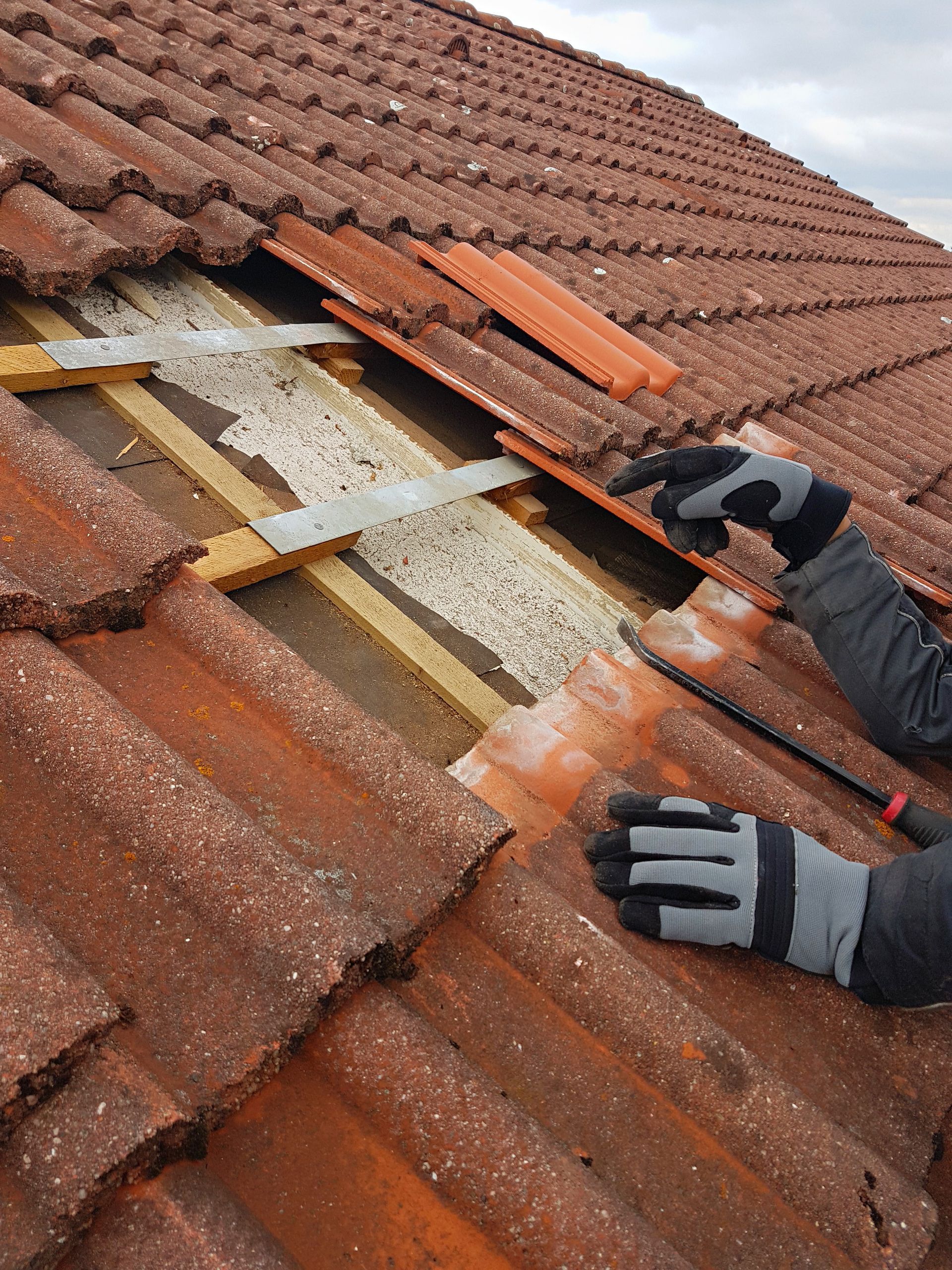 Worker repairing red tile roof, removing tiles to expose wooden battens and insulation under overcast skies