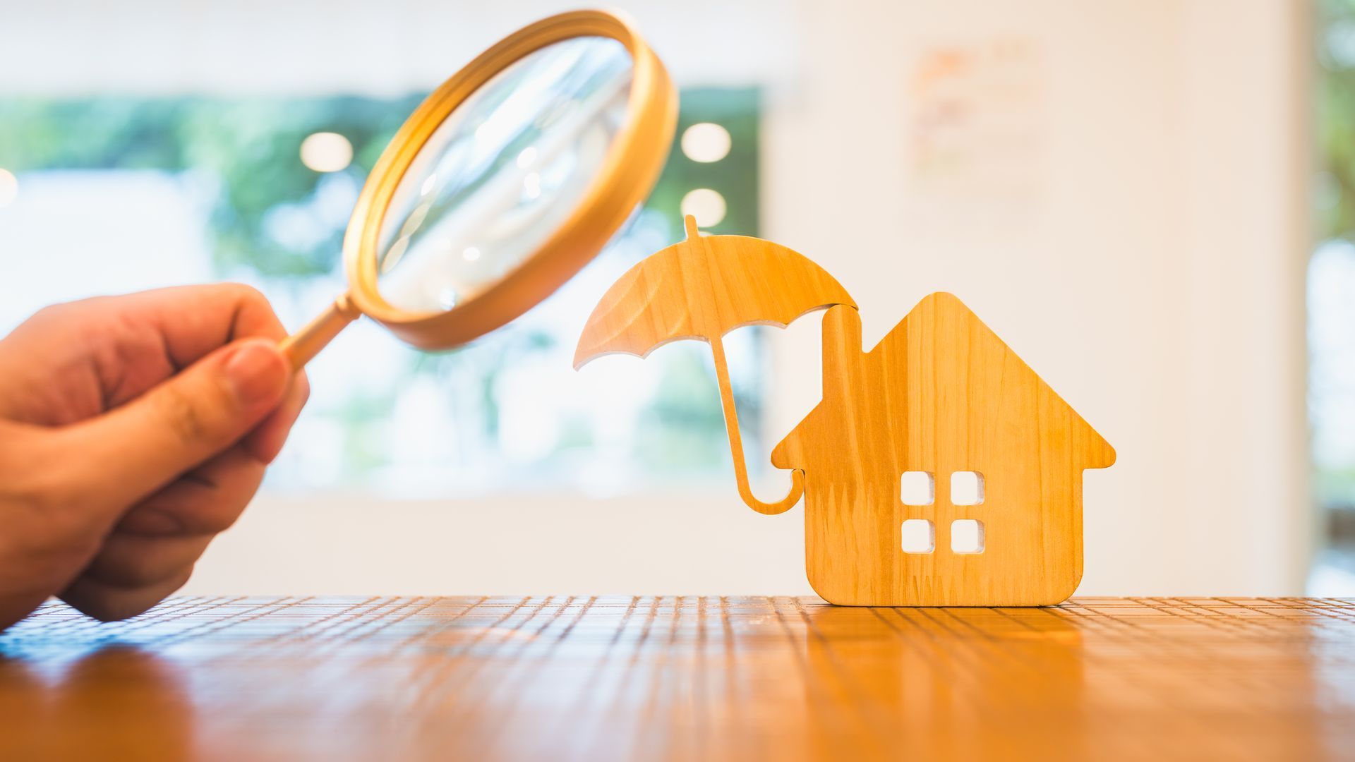 Hand holding magnifying glass over a small orange house with a broken umbrella, symbolizing home insurance inspection