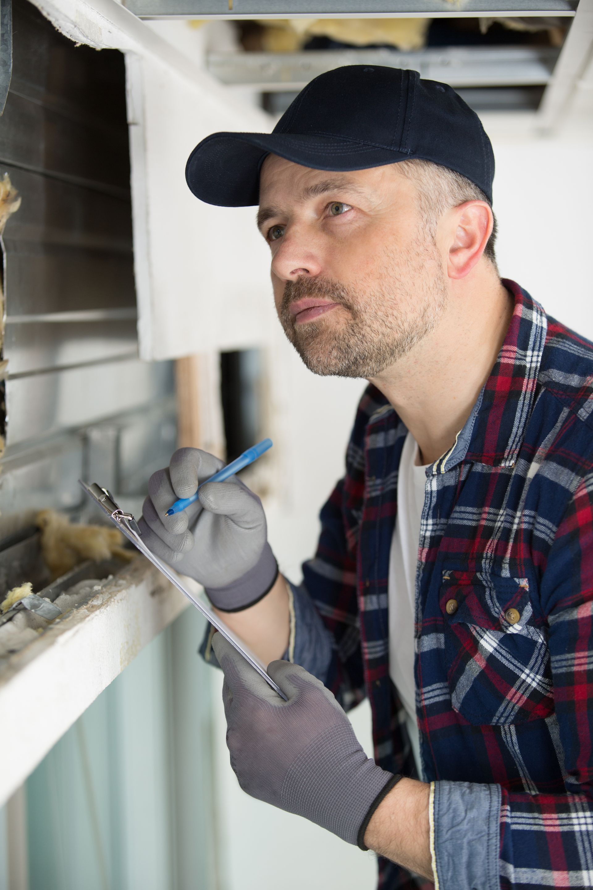 Worker in a black cap and gloves inspecting a vent opening with a flashlight.