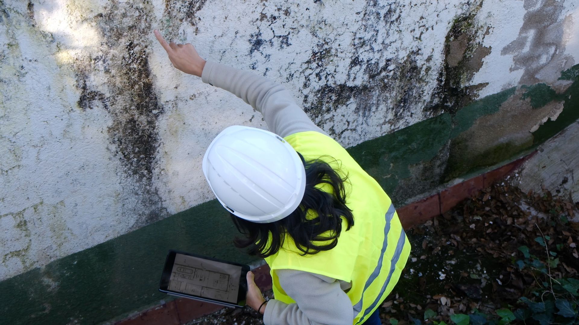 Worker in a white hard hat and yellow vest reaching up to inspect a stained wall outdoors.