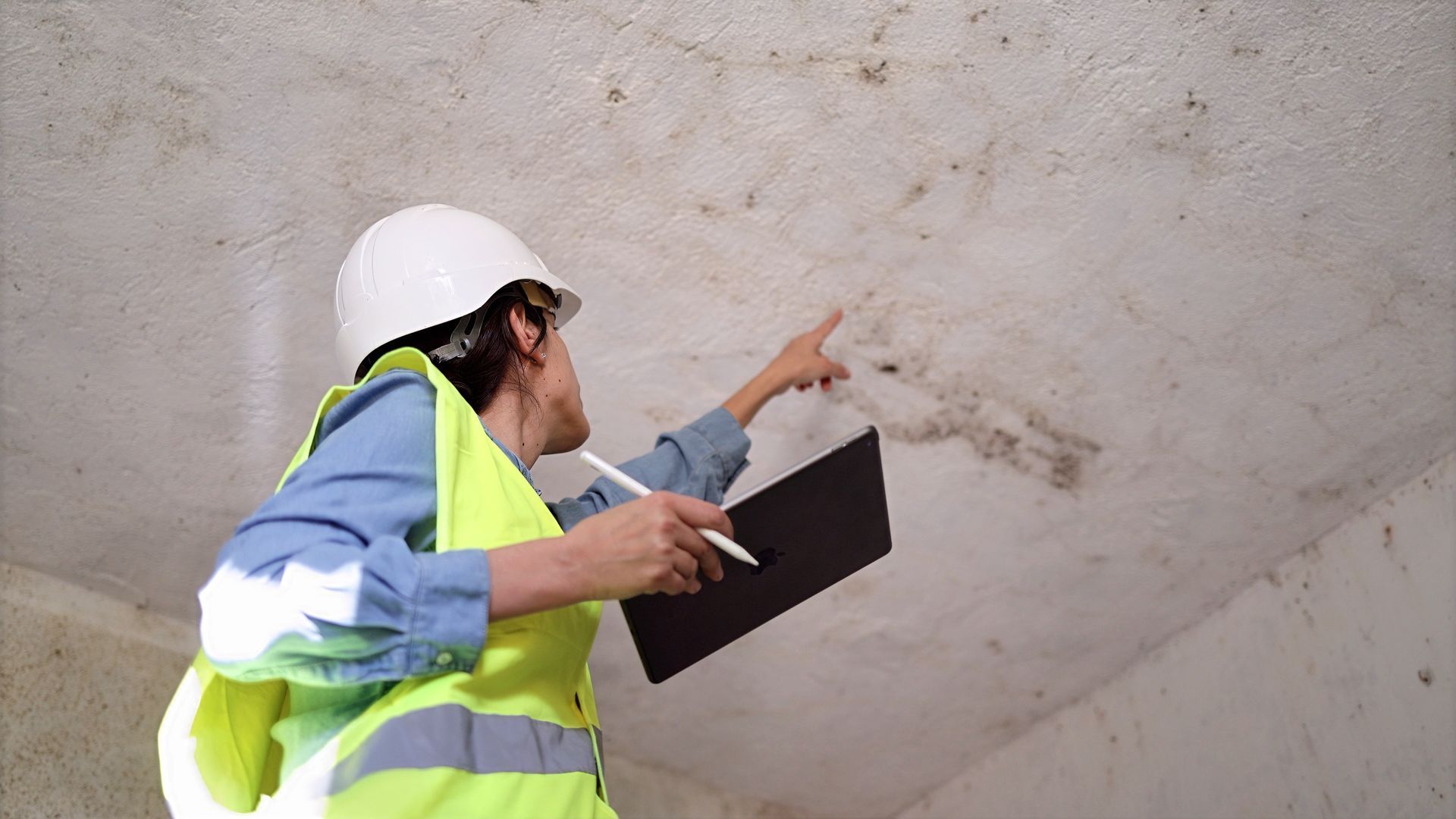 Worker in a hard hat and safety vest inspects a concrete wall with a clipboard.