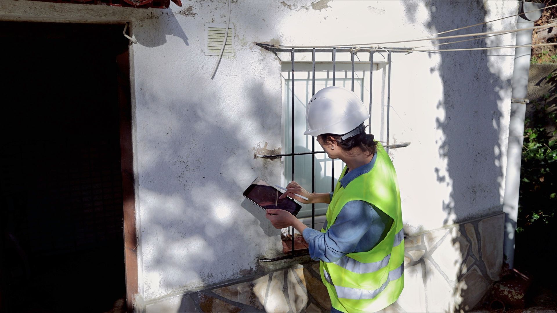 Worker in a hard hat and safety vest taking notes beside a sunlit building wall and window.