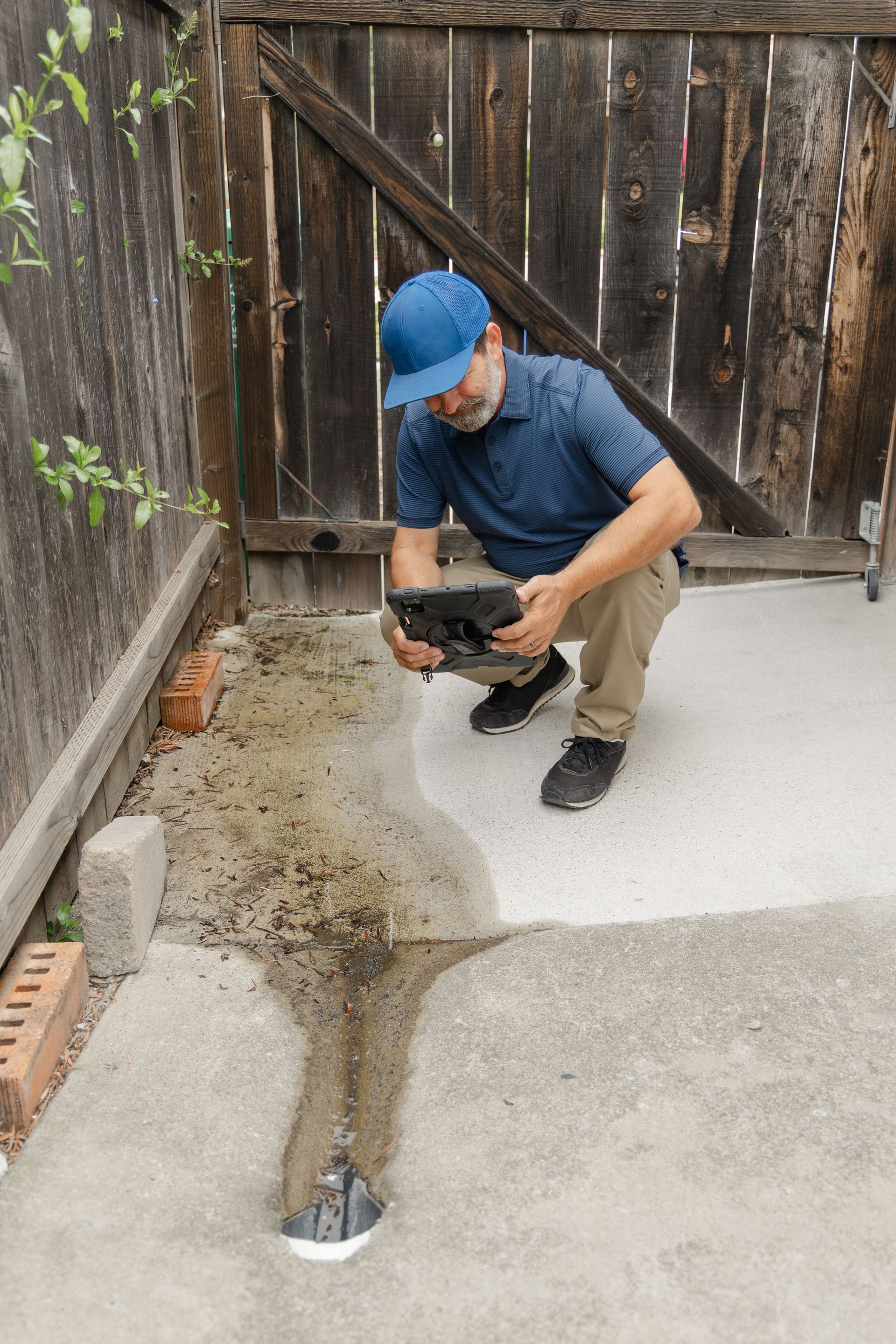 Worker crouching by a driveway drain, using a power tool to cut concrete near a wooden fence.