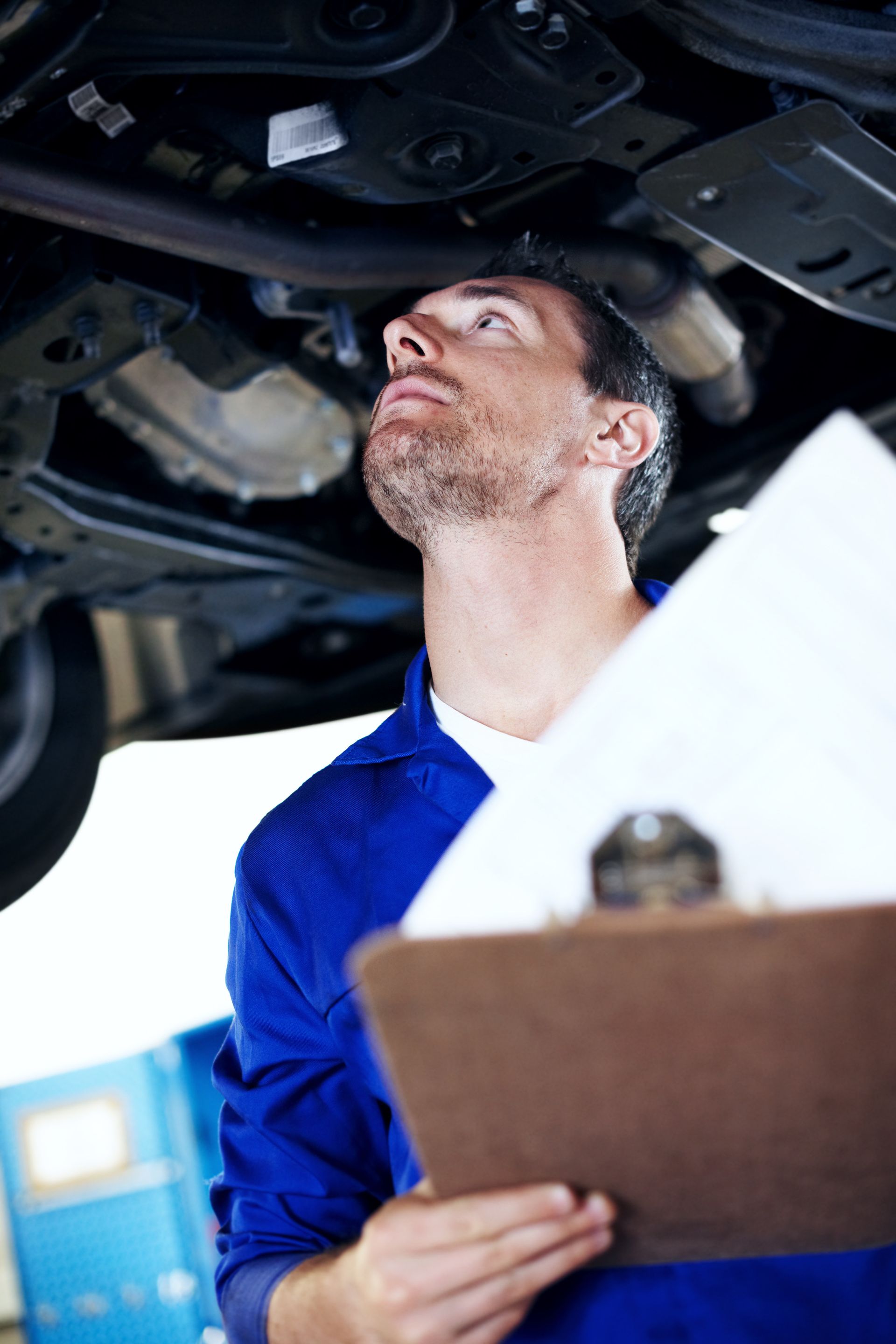 Mechanic in blue coveralls inspecting the underside of a car with a clipboard
