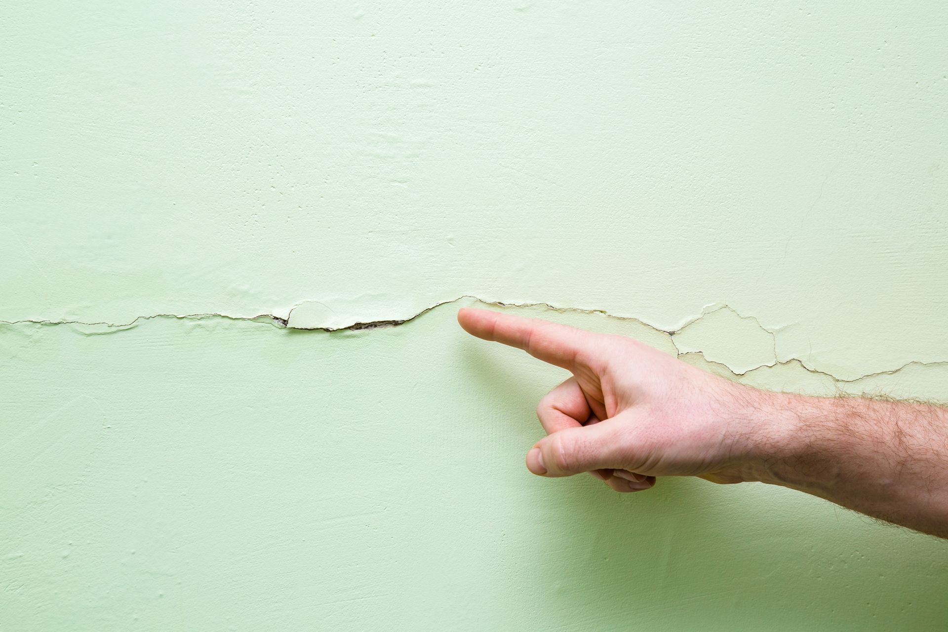 A hand pointing at a long crack in a pale green wall.