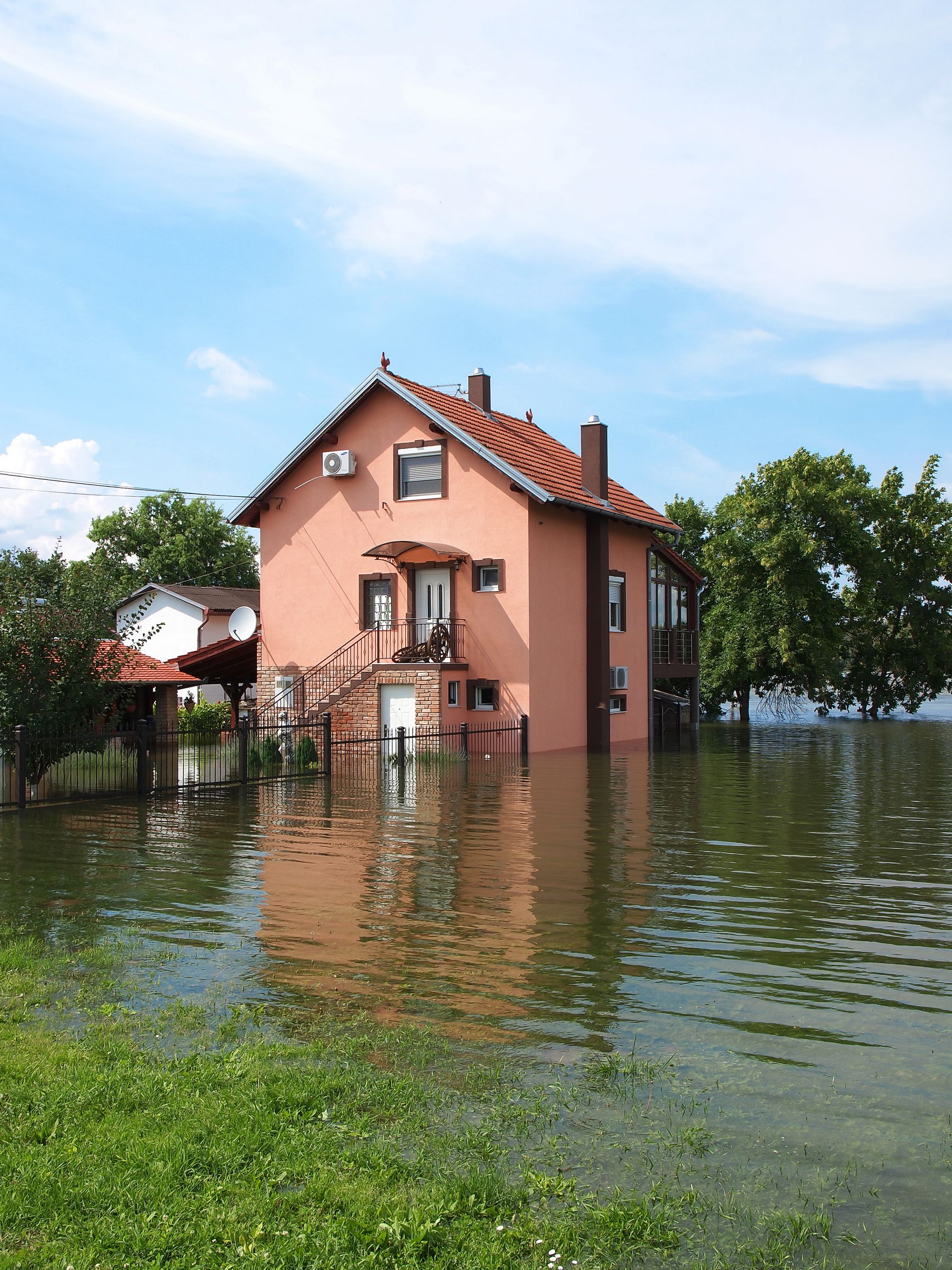 Pink house surrounded by floodwater beside a green lawn under a blue sky