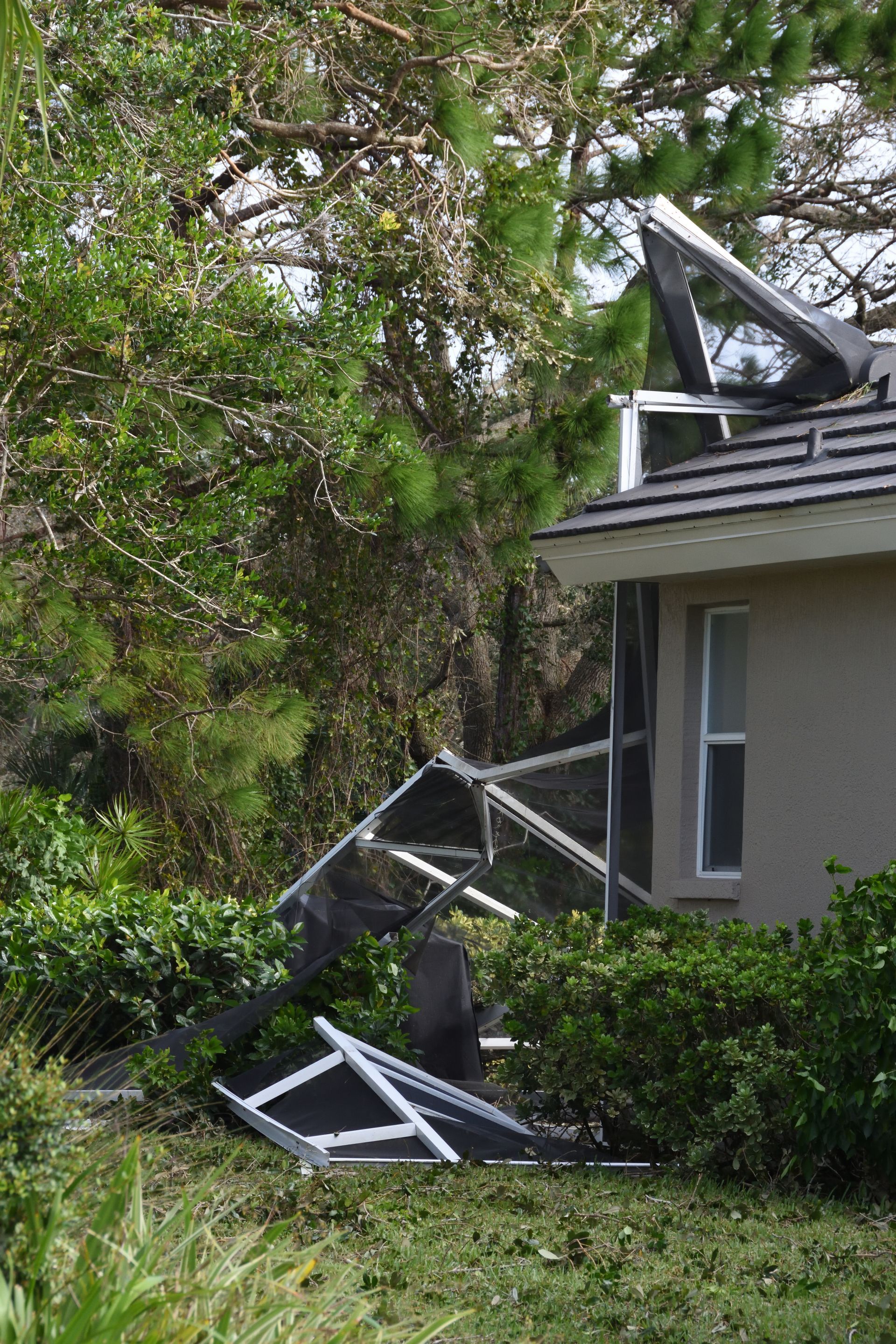 Collapsed metal structure beside a house, with debris in the yard and trees overhead.