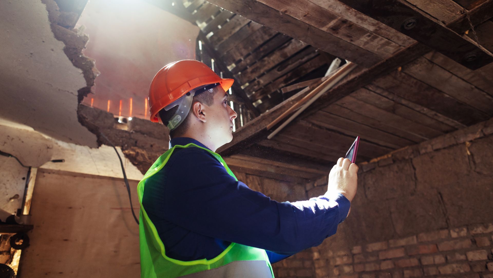 Worker in orange hard hat inspecting a damaged roof with sunlight shining through the opening