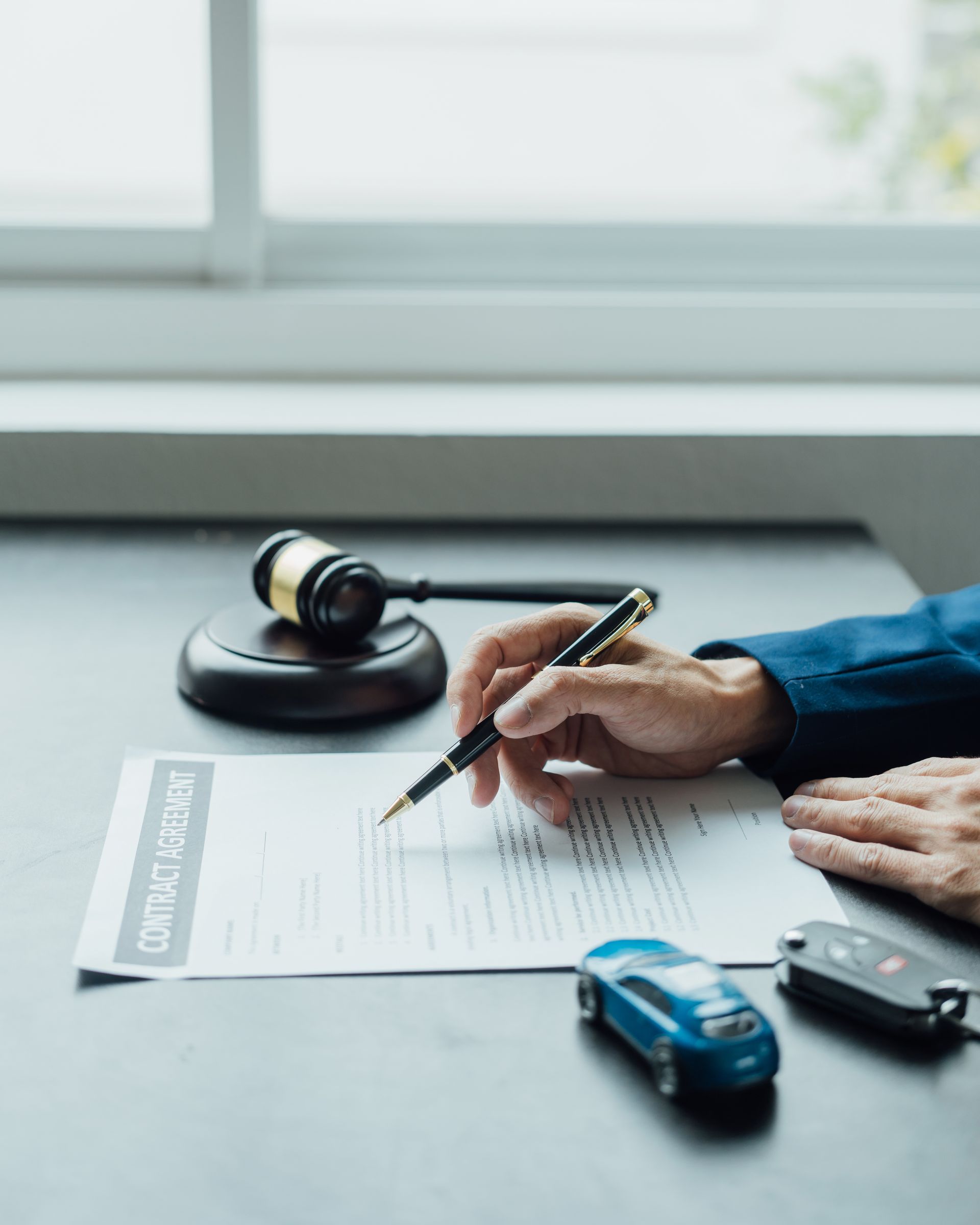 Hand signing a contract beside a gavel and toy cars on a desk