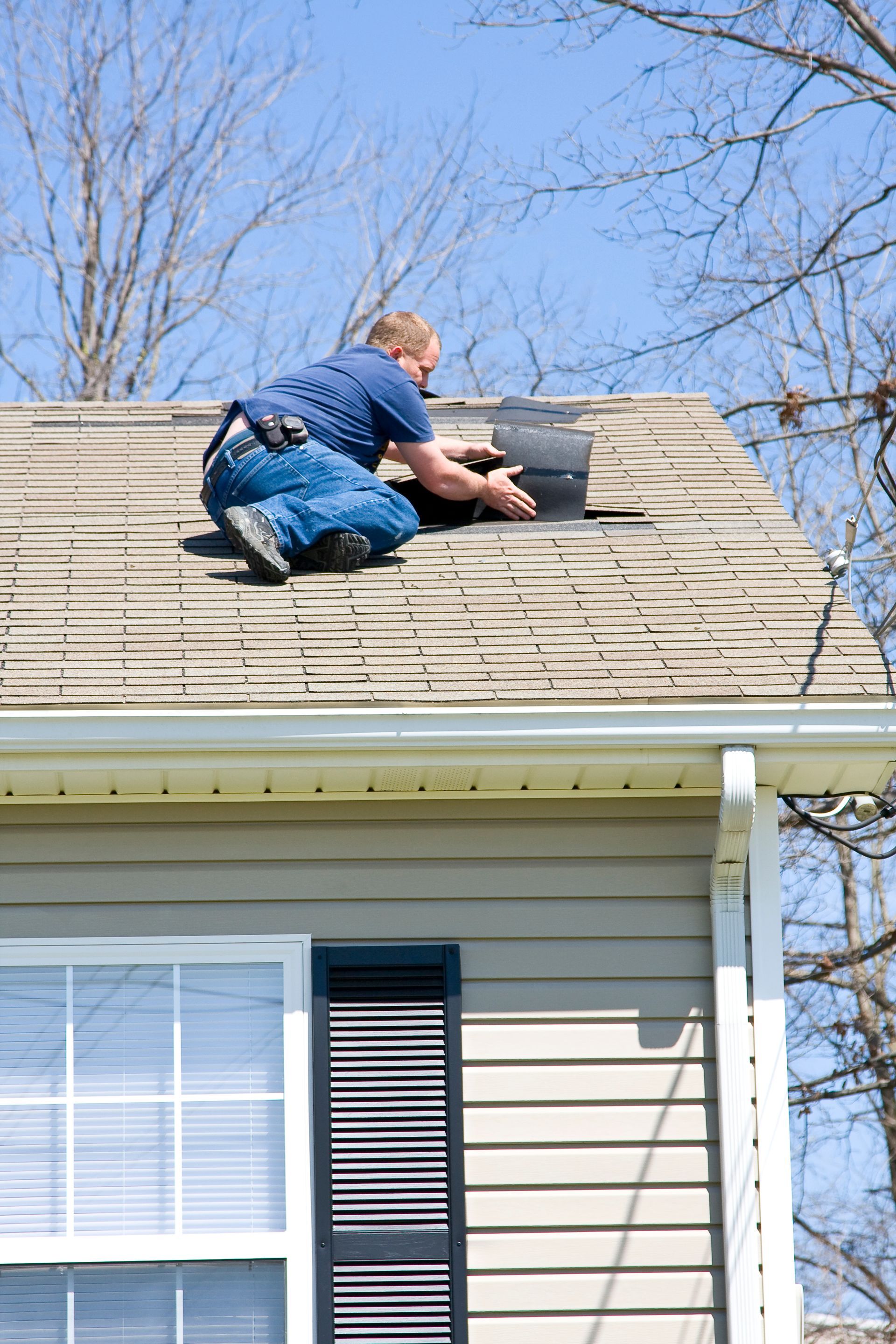 Worker repairing roof shingles on a house, kneeling near the ridge with tools.