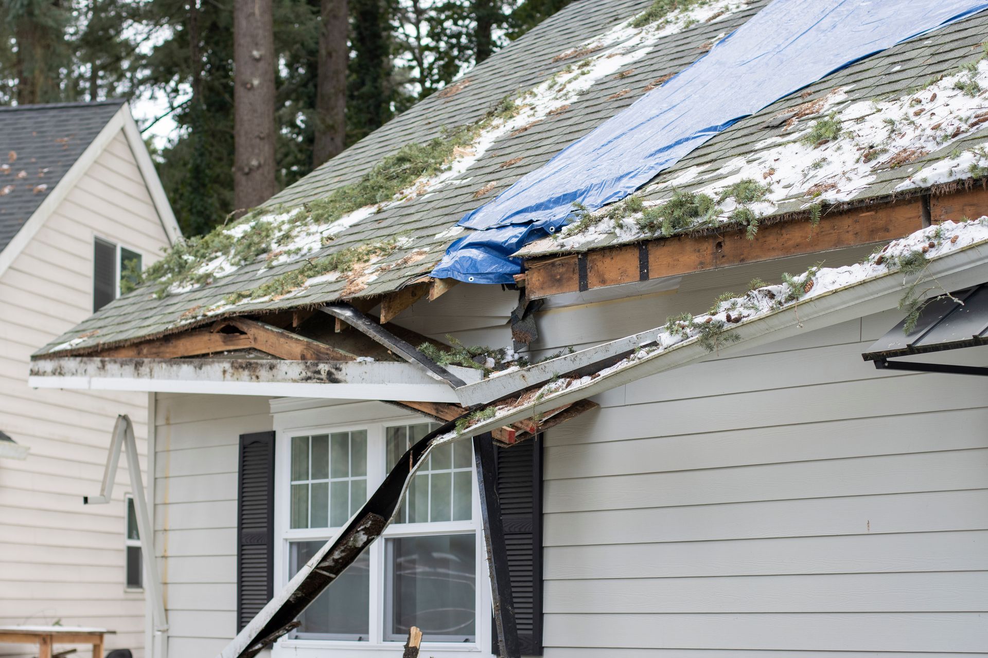 Damaged house roof with broken eaves and debris, partially covered in snow or tarp after storm damage