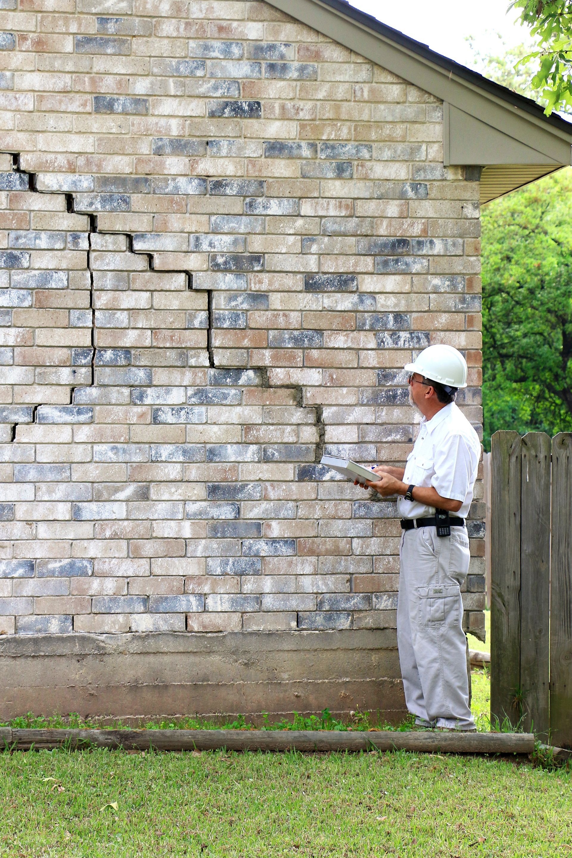 Worker inspecting cracked brick wall with a tool beside a house outdoors