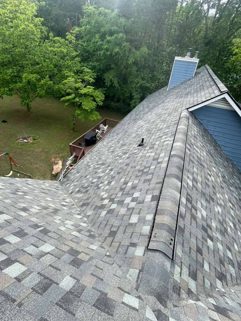 Roof shingles with a worker near the edge beside trees and a ladder.