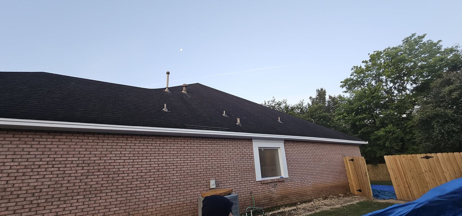 Brick house with dark roof and white window, viewed from a backyard under a clear sky