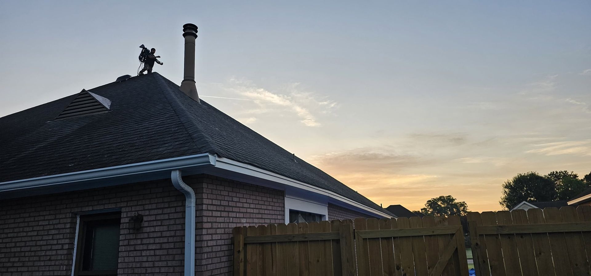 Brick house roof at sunset with chimney and wooden fence silhouetted against the sky