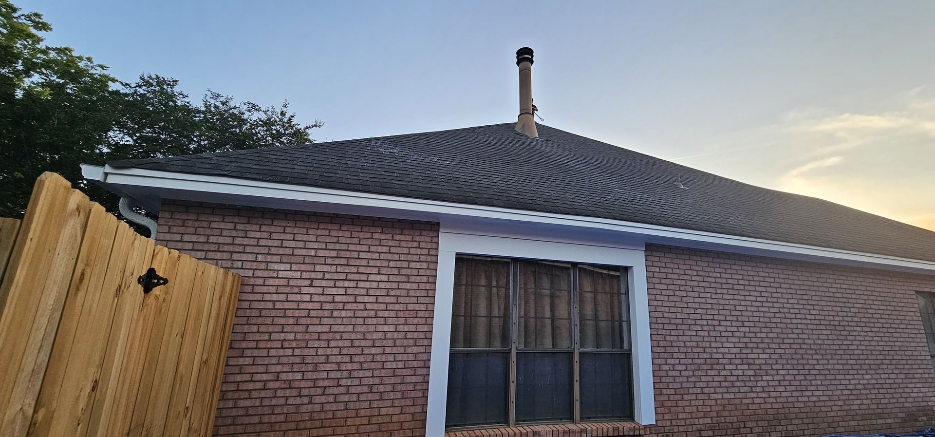 Brick house exterior with a dark shingled roof, window, and brick chimney at sunset