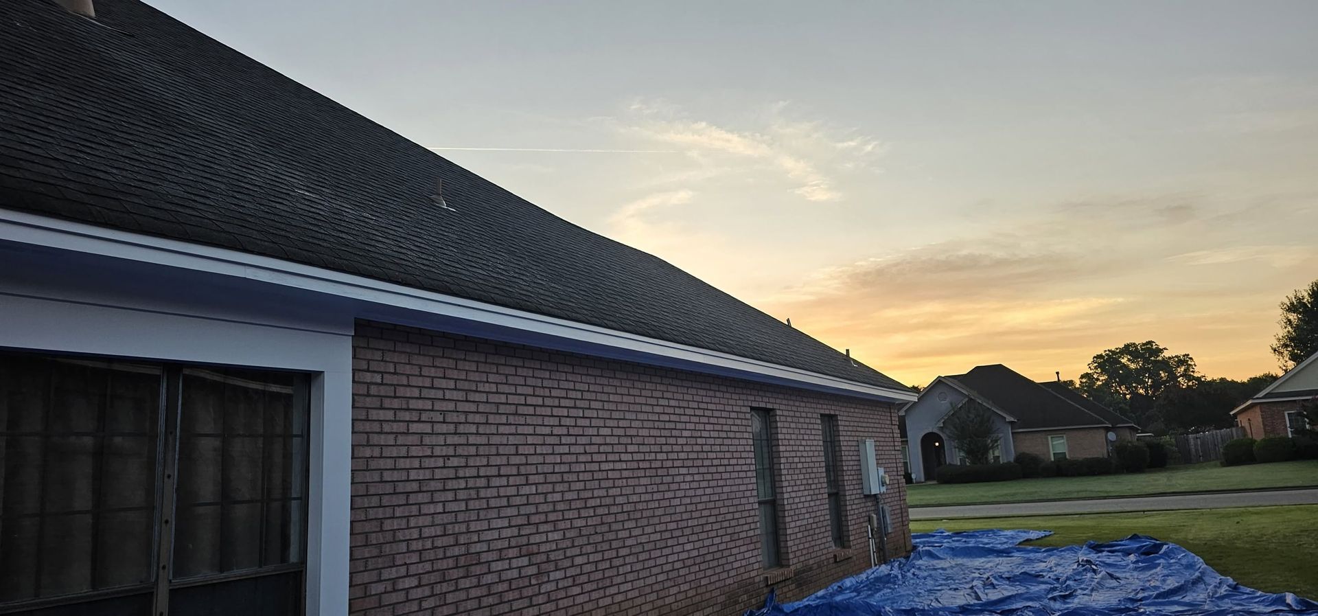Brick house at sunset with a dark roof and blue tarp-covered ground nearby