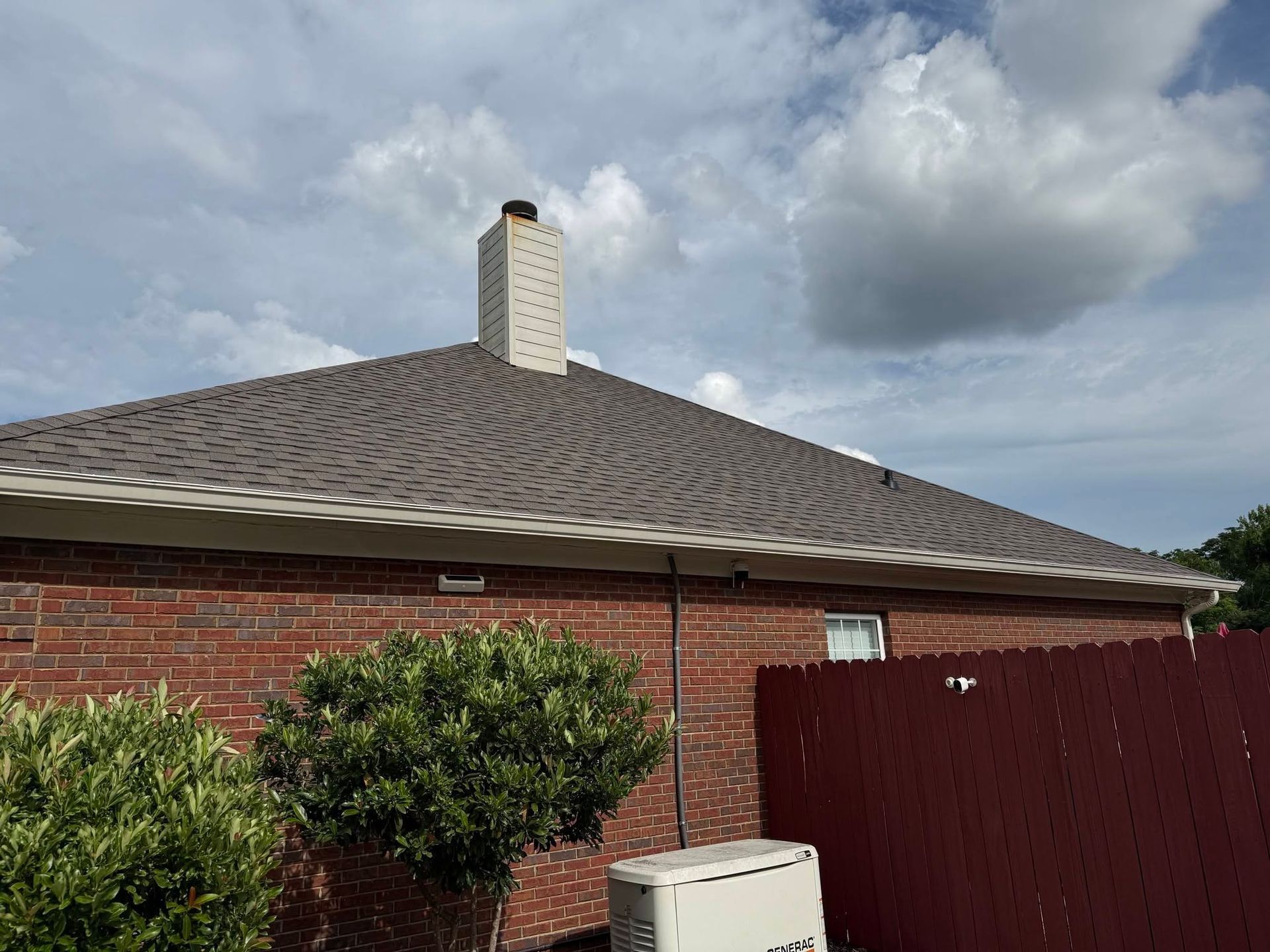 Red brick house with gray shingle roof, white chimney, and cloudy sky above a fenced yard