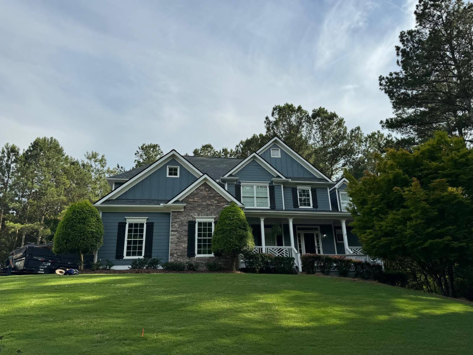 Gray two-story house with a porch on a grassy hill, surrounded by trees.