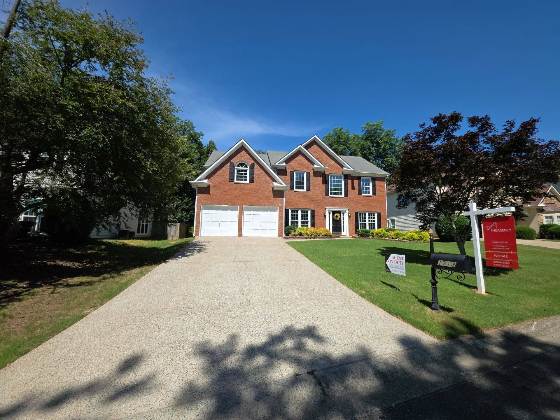 Brick suburban house with two garages, gravel driveway, and red real estate sign.