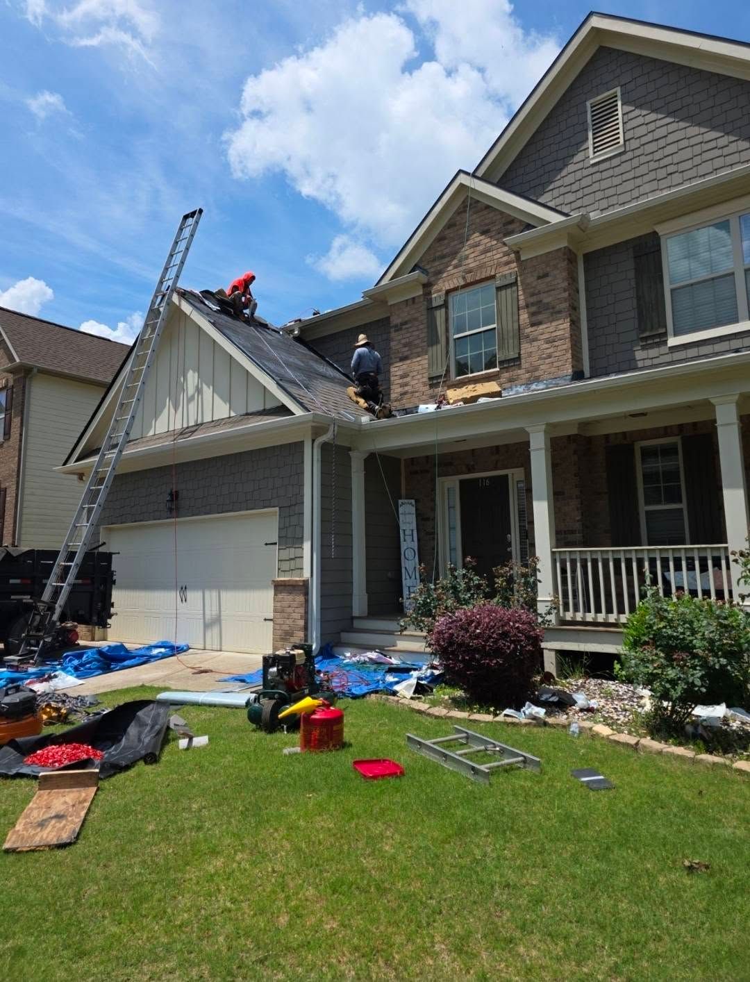 Workers repairing a two-story house roof and siding, with ladders and tools on the front lawn