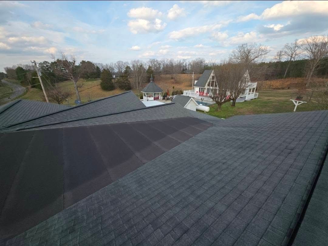 Roof with dark shingles overlooking houses and trees under a cloudy sky