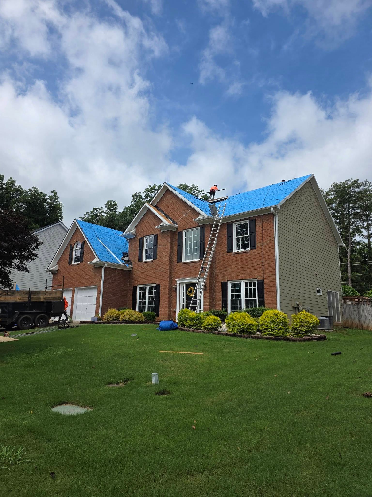 Brick house with blue roof under a partly cloudy sky, fronted by a green lawn and shrubs