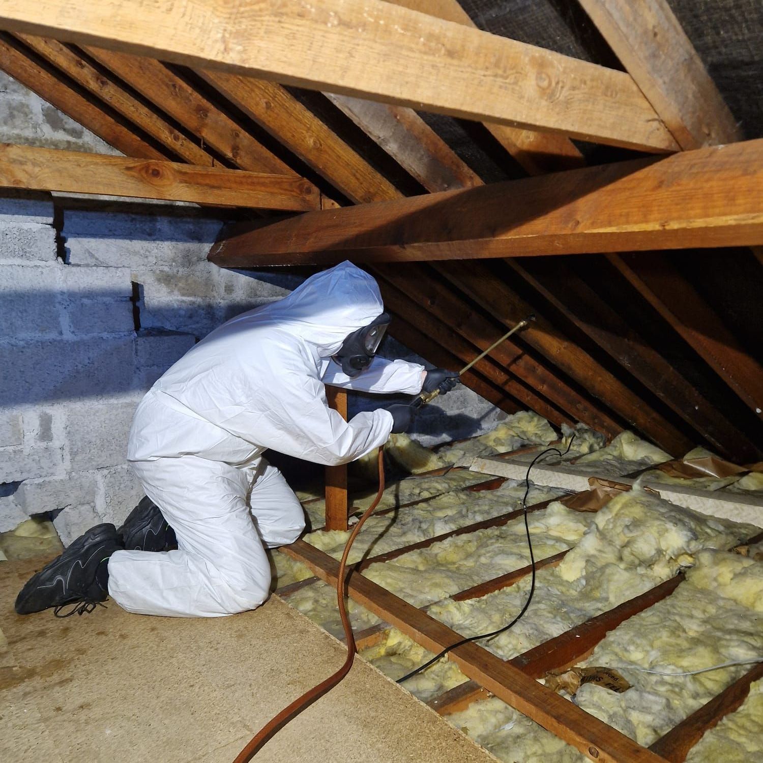 Person in protective suit spraying an attic's wooden beams, with insulation visible.