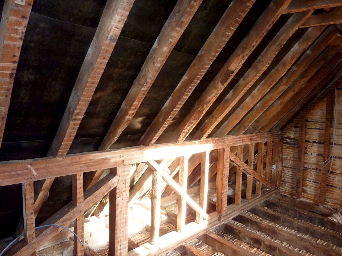 Interior view of an attic with exposed wooden beams and rafters. A railing is visible in the foreground.