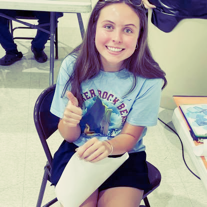 A person with long brown hair smiles while sitting on a chair, giving a thumbs up and holding a white paper.