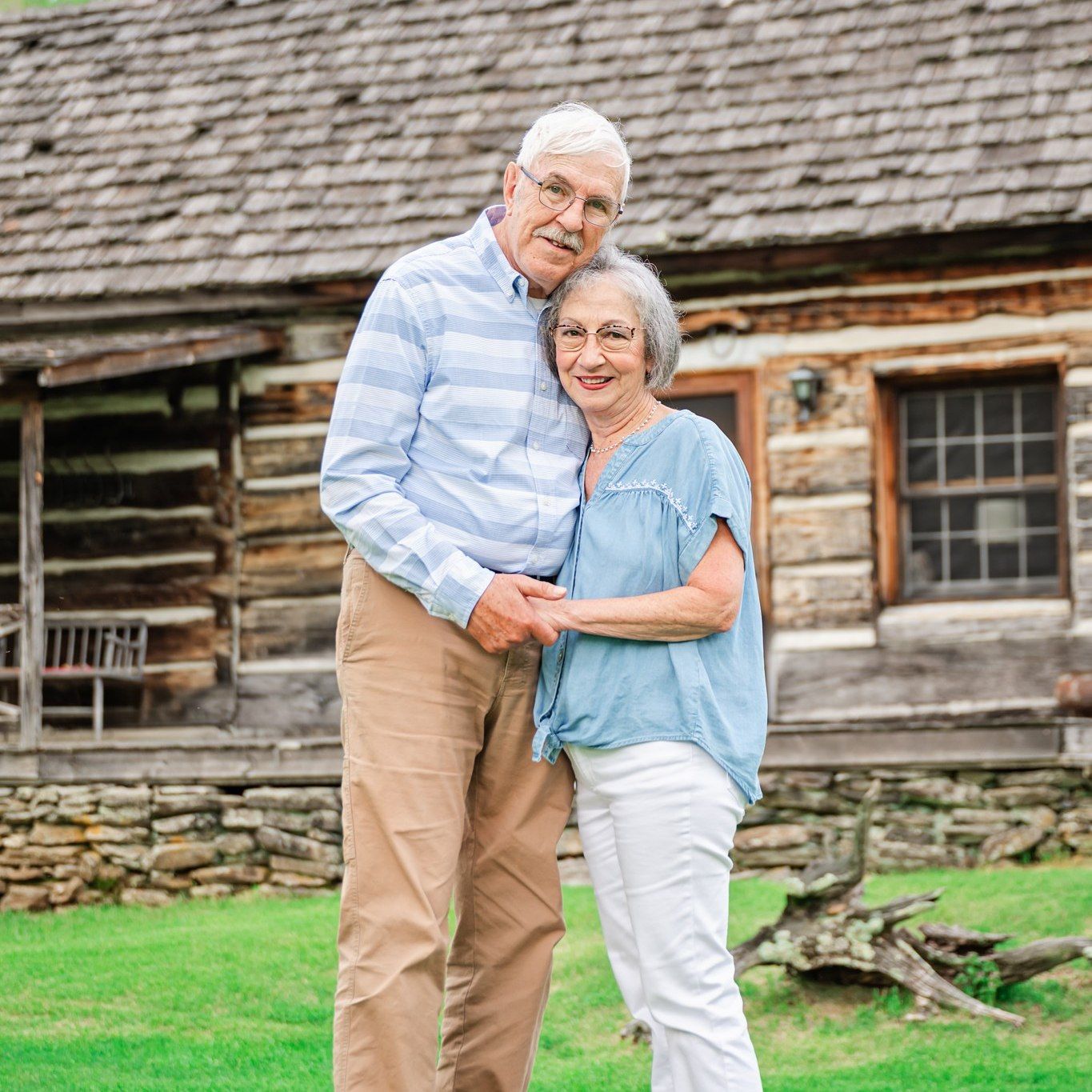 An older couple embracing while standing on the grass in front of a rustic wooden cabin.