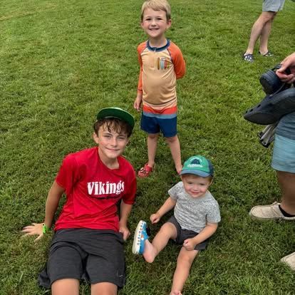 Three boys sit and stand on a grassy field; the older one wears a red shirt, while the younger two wear hats and play.
