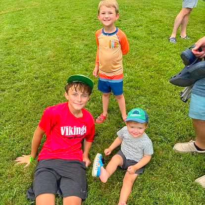 Three children sit or stand on a green lawn, smiling. One wears a red 