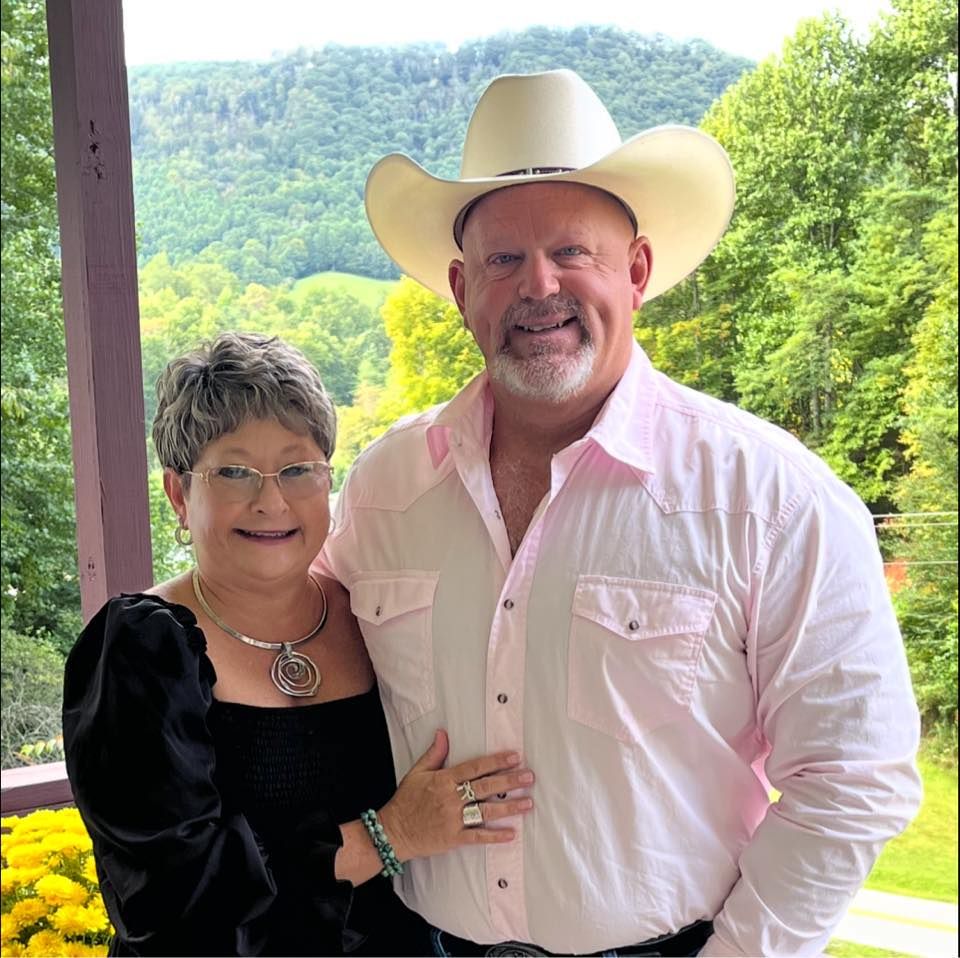 A smiling couple standing on a porch with green hills in the background. The man wears a cowboy hat and pink shirt.