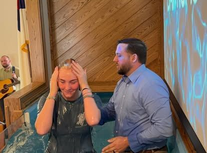 A man in a blue shirt performs a baptism for a person in a pool, while a musician plays guitar in the background.