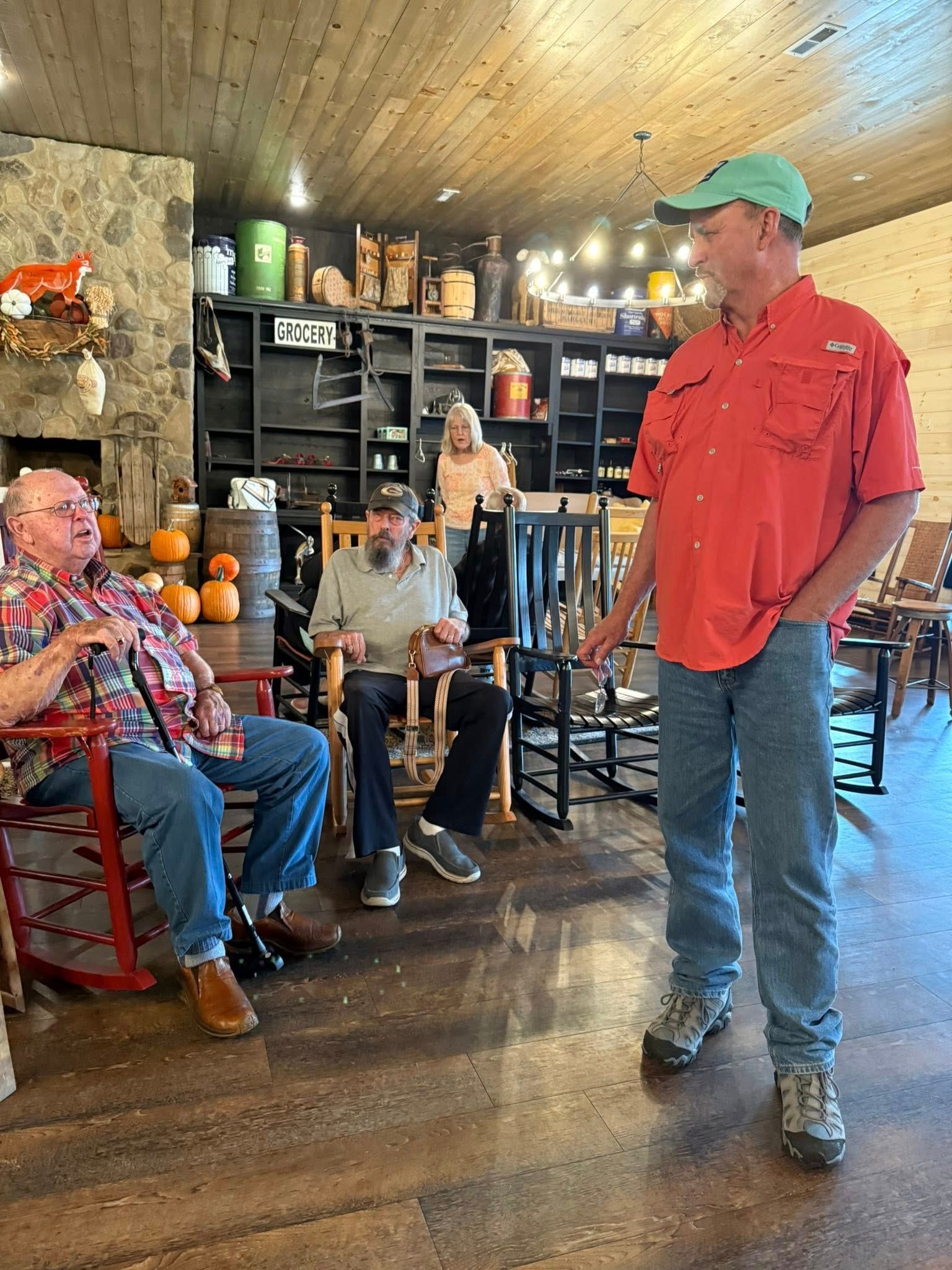 Three people in a rustic room with wooden walls and shelving; two sit in rocking chairs while one stands nearby.