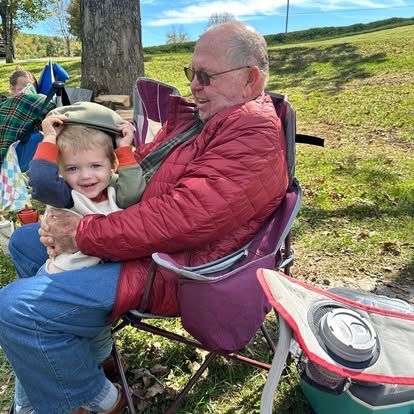 A person holding a young child in their lap, both smiling, sitting in folding chairs outdoors on a sunny day.