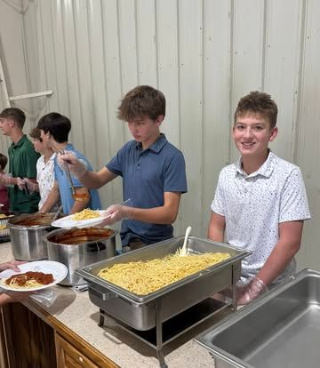 People serve spaghetti and sauce from large metal containers at a buffet-style meal in a room with paneled walls.