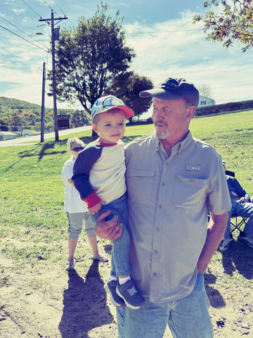 A person holds a young child in their arms while standing in a grassy area with a power line and trees in the background.
