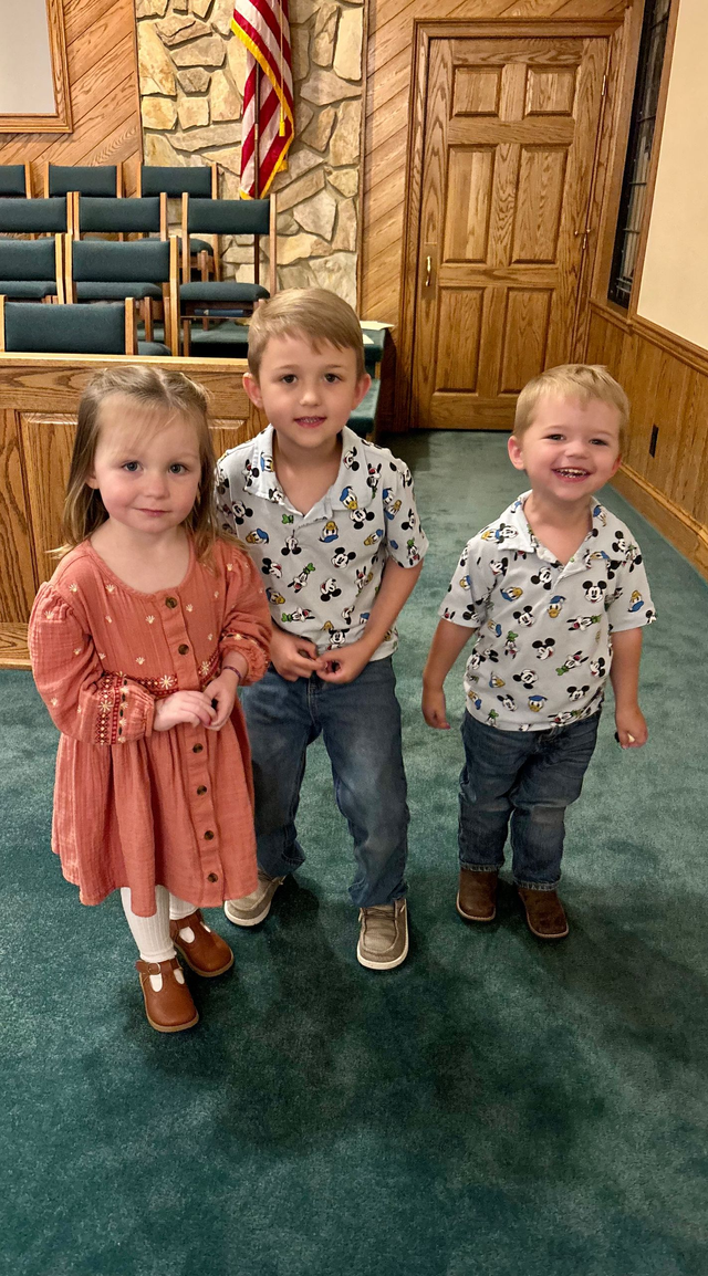 Three children stand smiling together in a room with wood-paneled walls, chairs, and a carpeted floor.