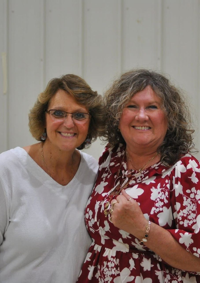 Two smiling people pose for a photo. One wears a white top and glasses, the other a burgundy and white floral blouse.