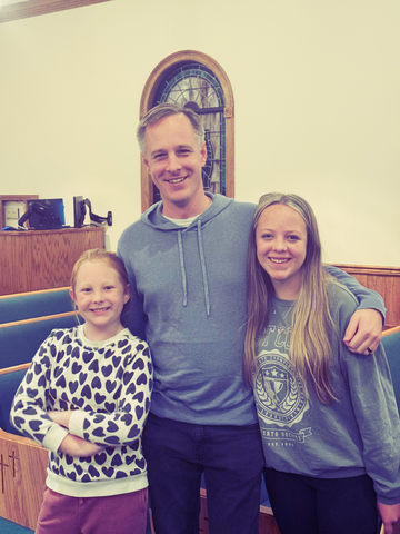 A man stands in a church between two smiling people with arms around their shoulders, wearing casual sweaters.