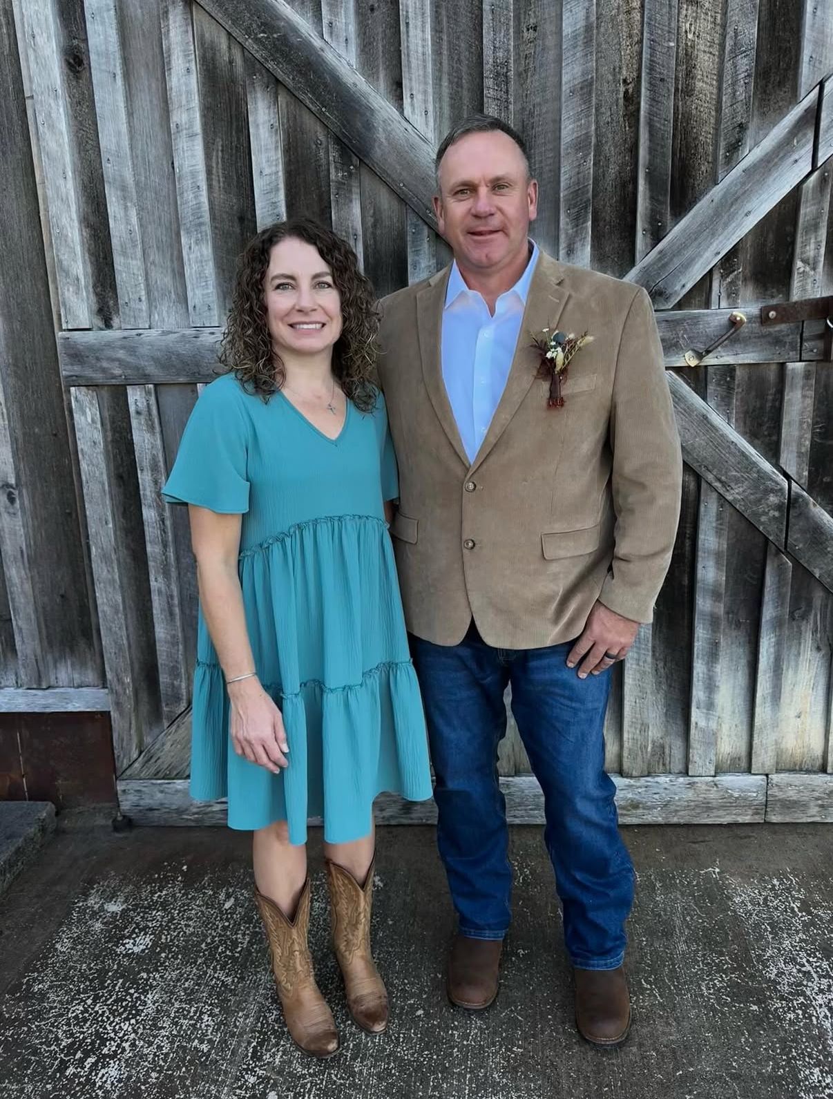 A smiling pair posing in front of a rustic wooden barn door; the woman wears a teal dress, the man a tan blazer and jeans.