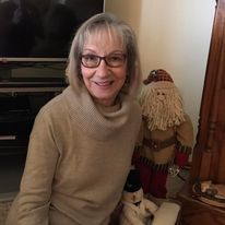 A person with shoulder-length gray hair and glasses in a tan turtleneck sweater, sitting near a decorative doll.
