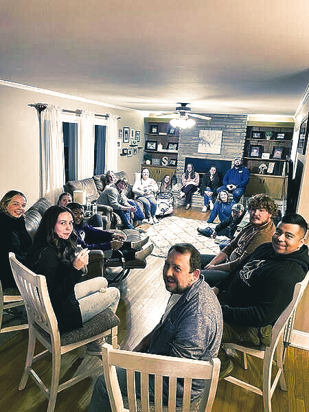 A large group of people sit together in a dimly lit, cozy living room, engaged in conversation during an evening gathering.